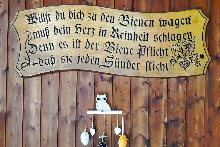 Geschnitzte Holztafel mit dem Imkerspruch an der Bretterwand des Bienenhauses, darunter ein kleines Windspiel