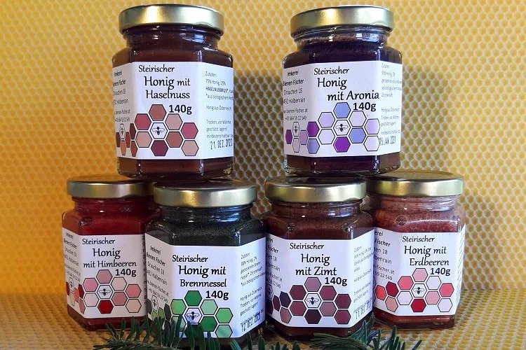 Sechs Waben-Sechskantgläser der Reihe Honig mit Zutat nebeneinander, die Etiketten nennen Haselnuss, Aronia, Himbeeren, Brennnessel, Zimt und Erdbeeren